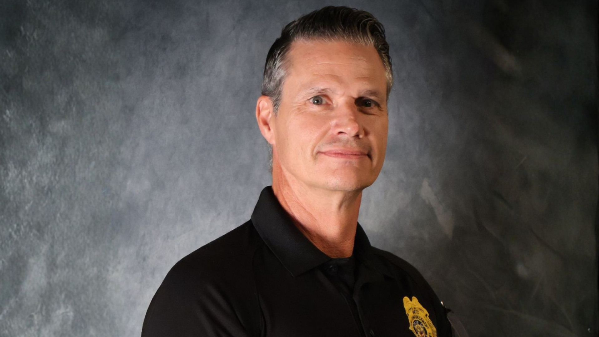 A man with gray hair in a black polo shirt bearing a law enforcement badge patch poses for a studio portrait against a gray backdrop.